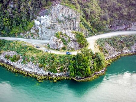 Eine kurvige Straße, die sich an steilen, felsigen Klippen entlang windet. Fahrzeuge sind auf einem kleinen, unbefestigten Parkplatz am Rande des Fjords abgestellt, umgeben von dichtem Grün und steinigen Ufern am türkisfarbenen ruhigen Fjord.