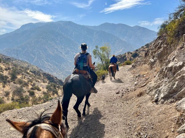 Reiter folgen einem schmalen Pfad in den Anden nahe Santiago de Chile. Die Szene zeigt eine Gruppe auf Pferden, eingebettet in eine trockene, bergige Landschaft mit Blick auf hohe Berge im Hintergrund.