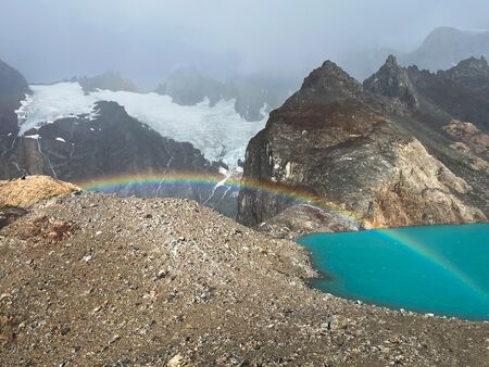 Ein Regenbogen, der in den Bergen von Patagonien an einem türkis blauen See unterhalb einer Gletscherlandschaft strahlt. Das Wetter im Hintergrund ist diesig. Der Vordergrund sonnig.