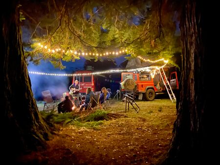 Mehrere Vans an die Lichterketten zur warmen Beleuchtung angebracht wurden, stehen zwischen Bäumen in der Natur. Es ist dunkel, ein paar Menschen sitzen um ein Lagerfeuer herum. Genau so stellt man sich das Vanlife vor.