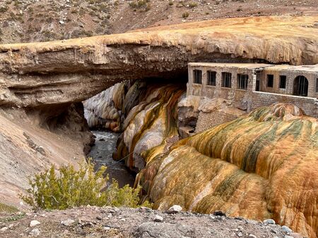 Die historische Puente del Inca, eine natürliche Felsbrücke mit bunten mineralischen Ablagerungen, überquert einen Fluss in einer trockenen Berglandschaft.