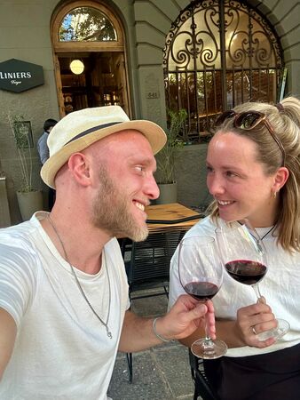 Ein fröhliches Paar sitzt draußen in einem Straßencafé in Mendoza, hebt die Weingläser zum Anstoßen, umgeben von einer charmanten städtischen Kulisse.