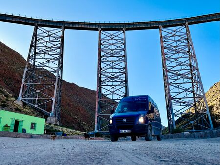 Ein blauer Van parkt unter dem hohen Viadukt des Tren a las Nubes in Argentinien, umgeben von roten Felsen und einem strahlend blauen Himmel. Im Vordergrund sind Hunde und ein kleines grünes Gebäude zu sehen.