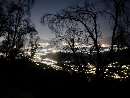 Nachtblick auf das beleuchtete Santiago de Chile, eingerahmt von dunklen Silhouetten kahler Bäume. Die Lichter der Stadt funkeln unter einem klaren, sternbesetzten Himmel.