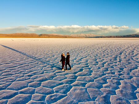 Zwei Personen laufen Hand in Hand über die weiten Salzflächen der Salinas Grandes in Argentinien, während die tiefstehende Sonne lange Schatten über die rissige Salzoberfläche wirft. Im Hintergrund sind Berge unter einem klaren blauen Himmel zu sehen.
