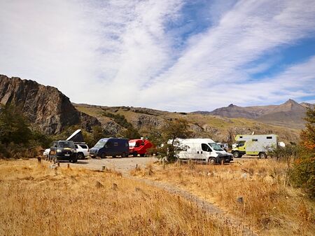 Mehrere Wohnmobile und Vans parken auf einem Stellplatz in El Chaltén. Der Platz ist umgeben von Felsen und Bergen, die im Hintergrund unter einem teilweise bewölkten Himmel zu sehen sind. Das Gelände ist von trockenen Gräsern bedeckt.