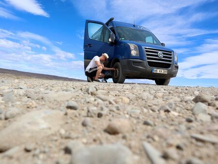 Ein Mann kniet neben einem blauen Volkswagen-Van auf der Ruta 40 in Patagonien und überprüft einen Reifen. Der Van steht auf einer steinigen Fläche unter einem blauen Himmel mit vereinzelten Wolken. Im Hintergrund die weite, trockene Landschaft.