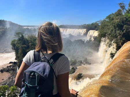 Eine Frau mit Rucksack steht auf einem Aussichtspunkt und blickt auf die majestätischen Iguazu Wasserfälle auf der argentinischen Seite. Die Wasserfälle stürzen kraftvoll herab, umgeben von üppiger Vegetation und einem klaren blauen Himmel.
