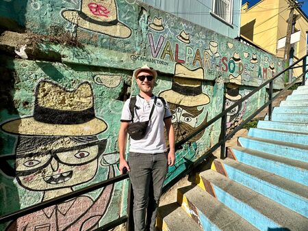 Ein Mann steht neben einer farbenfrohen, mit Graffiti bedeckten Treppe in Valparaíso, Chile. Die Treppe zeigt kunstvolle Darstellungen von Hüten mit der Aufschrift “Valparaíso de mi amor”.
