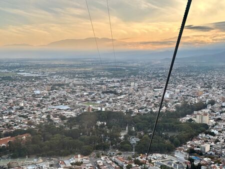 Ein Panoramablick auf die Stadt Salta, Argentinien, bei Sonnenuntergang, aufgenommen von einer Seilbahn. Im Hintergrund sind Berge und ein farbenfroher Himmel zu sehen.