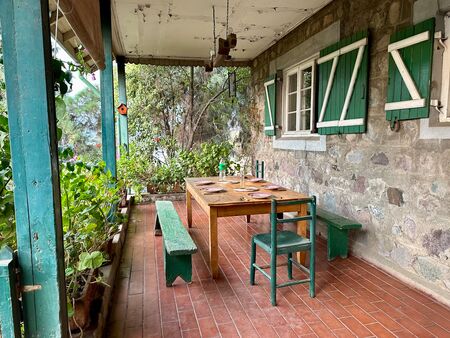 Ein gemütlicher Frühstückstisch auf einer steinernen Veranda, umgeben von grünen Fensterläden und einer reichen Auswahl an Pflanzen in einem hängenden Garten. Ein ruhiger, einladender Ort zum Entspannen im Freien - einfach perfekt in der Natur.