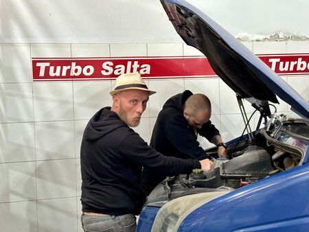 Zwei Männer arbeiten in einer Werkstatt in Salta, Argentinien, an einem blauen Fahrzeug, während einer humorvoll zur Kamera schaut. Im Hintergrund ist ein Schild mit der Aufschrift „Turbo Salta“ zu sehen.