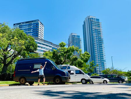 Zwei Vans auf einem Parkplatz in einer Großstadt (Buenos Aires). Ein junger Mann im Argentinien-Trikot steht vor dem blauen vorderen Van und streckt freudig die Arme in die Luft. So sieht Vanlife in Buenos Aires während der WM 2022 aus.
