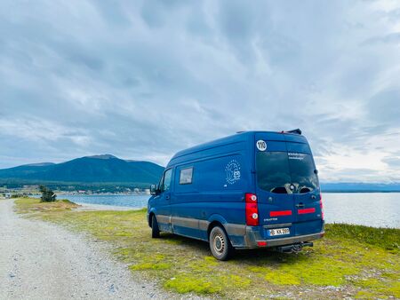 Ein blauer Van auf dem Grünstreifen neben einer Schotterstraße. Er parkt direkt am See, dem Lago Fagnano auf Feuerland. Im Hintergrund die Berge. Der Himmel ist bewölkt und etwas düster. Ein eindrucksvolles Foto.