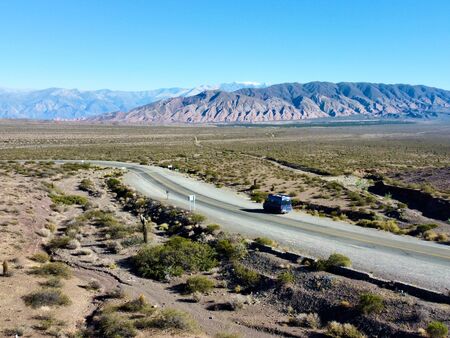 Ein blauer Van fährt auf einer abgelegenen Straße durch eine trockene Wüstenlandschaft, umgeben von Hügeln und Bergen, entlang der Ruta 40 in Argentinien unter einem klaren blauen Himmel.
