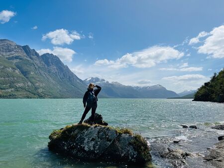 Eine Frau steht auf einem Felsen am Ufer des türkisfarbenen Lago del Desierto in Patagonien, umgeben von majestätischen Bergen und einem klaren, blauen Himmel mit weißen Wolken.