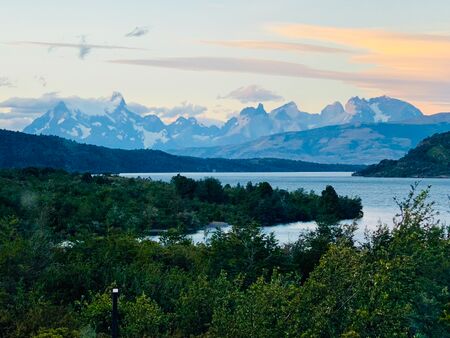 Das Bergpanorama Torres del Paine im Abendlicht. Der Himmel färbt sich rosa und im Vorderund ist ein bewaldeter Fluss zu sehen. Ein atemberaubender Anblicke.