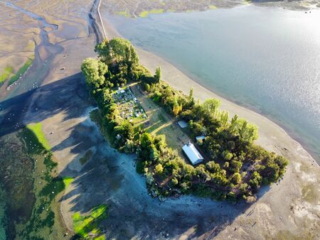 Eine Drohnenaufnahme der kleinen Isla Aucar auf Chiloé. Sie zeigt die Insel bei Ebbe, sodass viel Schlick, aber auch blaues Wasser drumherum ist. Das setzt die Bewaldung um die Kirche und den Friedhof der Insel herum gut in Szene. Es ist sonnig.
