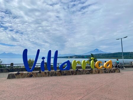 Der große blau- grün- gelbe Villarica- Stadtschriftzug. Im Hintergrund sind der See und rechts der Villarica Vulkan zu sehen. Es ist bewölkt, aber regnet nicht. Die Pflasterung der Promenade auf der das Schild steht ist rötlich.