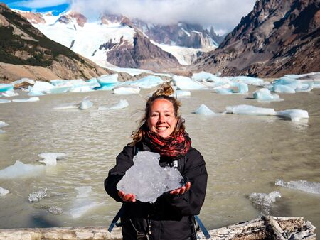 Eine Frau hält ein großes Stück Eis und lächelt in die Kamera, während sie vor der beeindruckenden Kulisse der Laguna Torre in El Chaltén steht. Im Hintergrund sind die verschneiten Berge und Gletscher zu sehen, während Eisstücke im Wasser treiben.