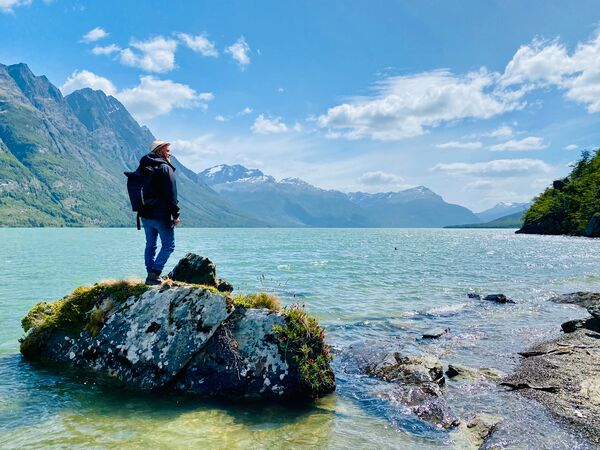 Ein Reisender steht auf einem Fels in einem türkisblauen See und schaut in die Ferne. Umliegend ein atemberaubendes Bergpanorama. Aufgenommen ist das Foto im Parque Nacional Tierra del Fuego.