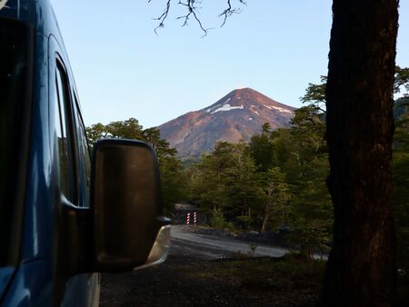 Linksseitig ein Teil eines blauen Vans inkl. Rückspiegel im Vordergrund und mittig der imposante im Abendlicht leuchtende aktive Villarica Vulkan. Ein wilder Stellplatz mit perfektem Blick.