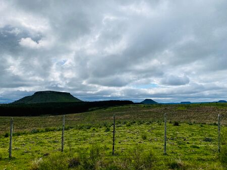 Flache Tafelberge erheben sich unter einer bewölkten Himmel im Valle de Lunarejo, Uruguay. Im Vordergrund ist eine grüne Weidelandschaft zu sehen, die durch einen einfachen Drahtzaun abgegrenzt ist.