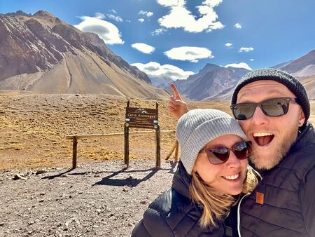 Ein lächelndes Paar in Sonnenbrillen und Mützen posiert vor einem Schild des Cerro Aconcagua, umgeben von beeindruckenden Berggipfeln und blauem Himmel.