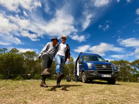 2 Wanderer, die ihre Füße mit Wanderschuhen an in die Kamera halten. Daneben ein blauer Van. Das Bild vermittelt Freude über die Wanderschuhe und Abenteuerlust. Der Himmel ist blau mit leicht windig wirkenden weißen Wolken.