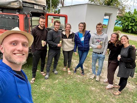 Eine Gruppe von acht jungen Reisenden posiert fröhlich mit Getränken in der Hand vor einem roten Geländewagen auf einem Campingplatz in Salta, Argentinien.