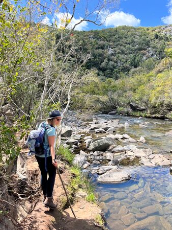 Eine Wanderin mit Rucksack und Wanderstock steht am Ufer eines klaren Flusses in der Quebrada de los Cuervos in Uruguay. Umgeben von dichtem Grün und Felsen, blickt sie auf das Wasser, während der Himmel strahlend blau ist.