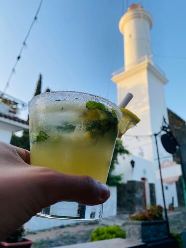 Eine Hand hält einen frisch zubereiteten Mojito-Cocktail und prostet damit dem im Hintergrund sichtbaren Leuchtturm von Colonia del Sacramento zu. Der markante Leuchtturm ragt majestätisch über der Szene empor.