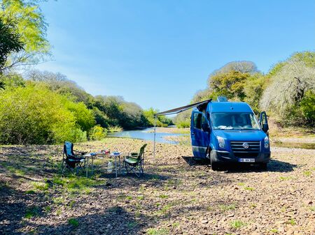 Ein blauer VW-Campervan steht auf einem felsigen Ufer neben einem Fluss, umgeben von üppigem grünem Laub und Bäumen. Vor dem Van sind zwei Campingstühle und ein Tisch mit Frühstücksutensilien aufgestellt. Der Himmel ist klar und blau.