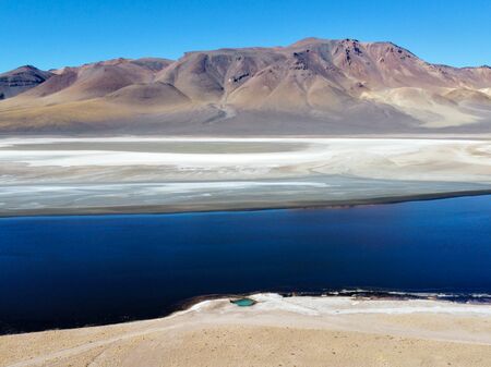 Eine ruhige, tiefblaue Lagune erstreckt sich vor trockenen, farbenfrohen Bergen in der abgelegenen Puna-Region Argentiniens unter einem klaren, blauen Himmel. Die Landschaft ist geprägt von Kontrasten zwischen Wasser, Salzflächen und kahlen Berghängen.