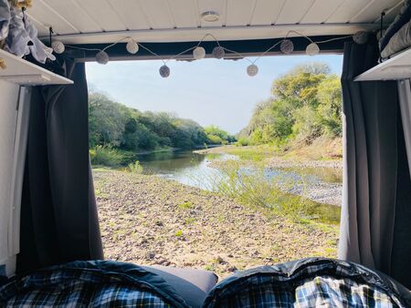Blick aus einem Campervan auf einen idyllischen Fluss im Valle de Lunarejo, Uruguay. Die Schotterbank am Flussufer ist von üppiger Vegetation umgeben, und der Himmel ist klar und blau.