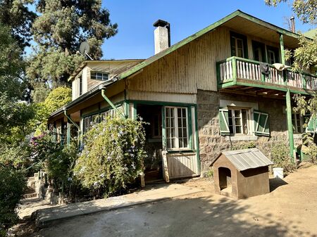 Ein zweistöckiges Landhaus mit grünem Holzrahmen, Holzbalkon, umgeben von Bäumen und Blumen, mit einer Hundehütte im Vordergrund.