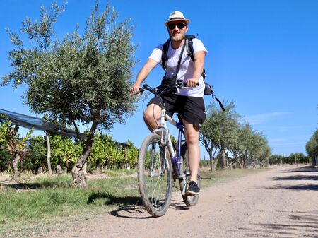 Ein Mann mit Sonnenhut und Rucksack fährt auf einem Fahrrad durch eine malerische Weinberglandschaft in Mendoza unter klarem, blauem Himmel.
