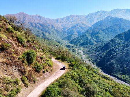 Ein Van fährt auf einer kurvigen Bergstraße, umgeben von dichter grüner Vegetation und majestätischen Bergen, in einer abgelegenen Gegend in Argentinien unter klarem, blauem Himmel.