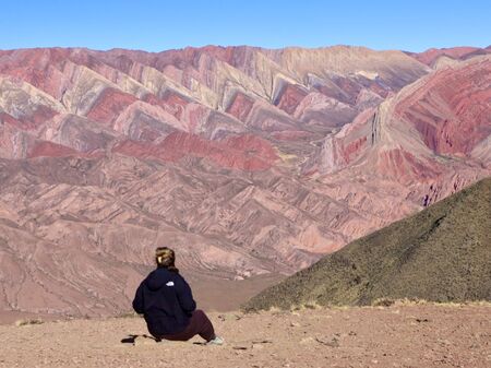 Eine Person in dunkler Kleidung sitzt auf dem Boden und schaut auf die beeindruckende, mehrfarbige Berglandschaft der Serranía de Hornocal in Argentinien, unter einem klaren blauen Himmel.