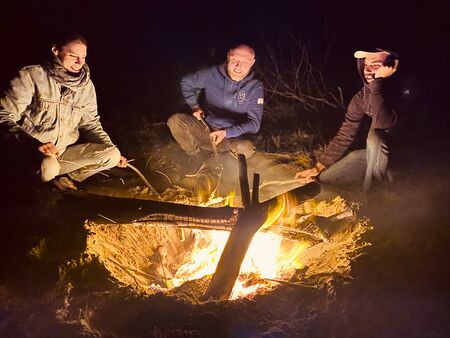 3 junge Erwachsene sitzen um eine Lagerfeuer und freuen sich über die ausstrahlende Wärme. Es sieht nach einem sehr gemütlichen Lagerfeuerabend aus.