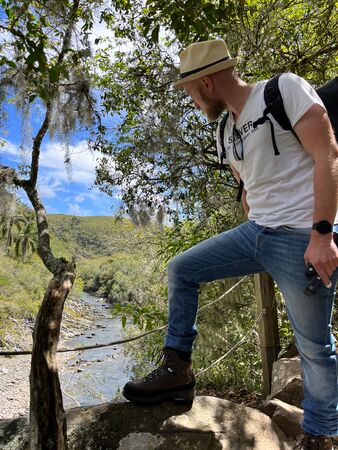 Ein Wanderer schaut in vegetationsreicher Landschaft hinunter zu einem Fluss. Sein Bein ist motiviert aufgestellt und er trägt einen Hut und Rucksack.