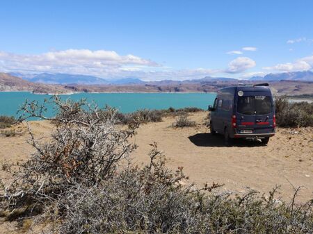 Ein blauer Van steht an einem türkisfarbenen See in Patagonien im Patagonia Nationalpark (Argentinien). Umgeben von trockenen Büschen, Sand und in der Ferne ein Bergpanorama. Der Himmel ist blau mit vereinzelten Wolken.