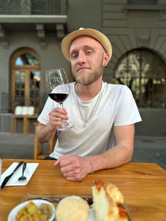 Ein Mann hält ein Rotweinglas am Esstisch hoch und ist bereit einen Malbec in Mendoza zu probieren. Er trägt einen Hut. Im Hintergrund ein schönes dunkelgraues Gebäude der Stadt.