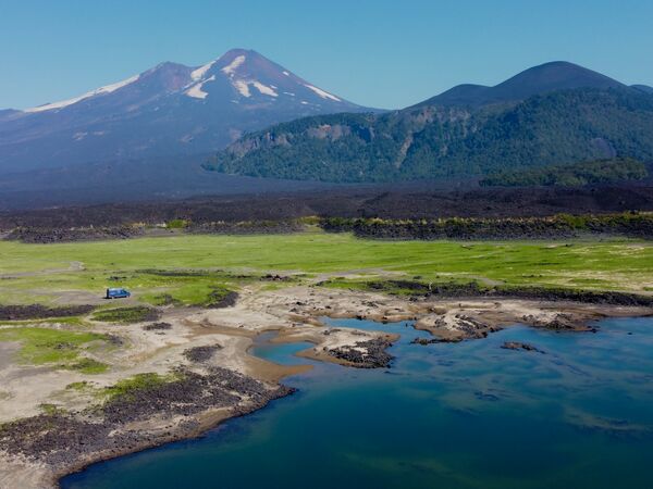 Ein blauer Campervan steht allein auf einer grünen Wiese im Conguillío-Nationalpark, umgeben von einer spektakulären Vulkanlandschaft. Im Hintergrund erhebt sich der schneebedeckte Llaima-Vulkan, flankiert von weiteren dunklen Vulkanhügeln.
