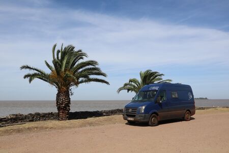 Unser blauer VW Crafter Wohnmobil steht an einer idyllischen Promenade in Colonia del Sacramento, Uruguay, unter zwei majestätischen Palmen. Die Promenade bietet einen malerischen Blick auf den Río de la Plata.