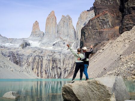 Zwei Personen stehen auf einem Felsen am Ufer eines türkisfarbenen Sees und blicken auf die majestätischen Torres del Paine Türme. Sie umarmen sich und winken, umgeben von beeindruckenden Felsformationen und klarem Himmel.