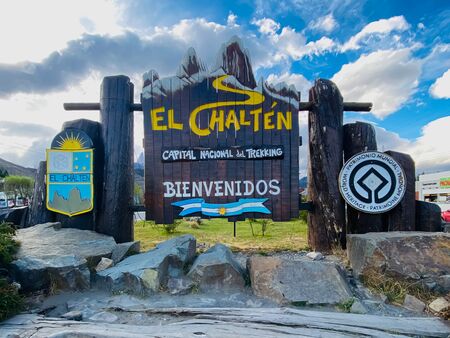 Das kunstvolle und eindrucksvolle hölzerne Ortsschild von El Chaltén. Der Himmel im Hintergrund ist leicht bewölkt, aber sonnig. Auf dem Schild steht u.a.  Capital Nacional del Trekking.