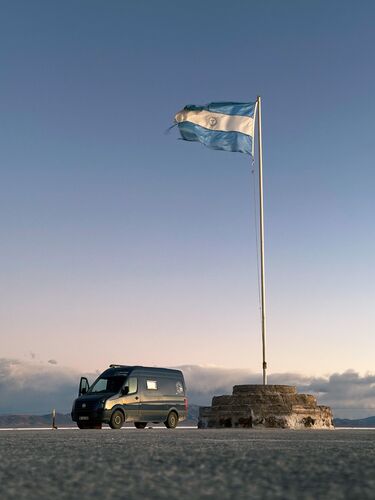 Ein blauer VW Crafter, der unter einer argentinischen großen Flagge auf den Salinas Grandes in Argentinien im romantischen Abendlicht steht. Der Himmel ist dabei blau bis leicht rosa gefärbt. Das Bild vermittelt einen romantischen Vanlife Moment.