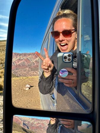 Eine lächelnde Frau mit Sonnenbrille hält zwei Finger in einem Friedenszeichen aus dem Fenster eines Vans, während sie im Seitenspiegel die farbenprächtige Berglandschaft von Serranía de Hornocal in Argentinien reflektiert sieht.