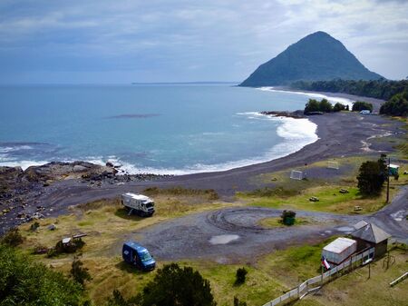Ein wilder Stellplatz an einem abgelegenen, schwarzen Sandstrand entlang der Carretera Austral in Chile. Zwei Fahrzeuge stehen auf einem grasbewachsenen Platz mit Blick auf das türkisfarbene Meer. Im Hintergrund liegen ein bewaldeter Hügel und die Küste.